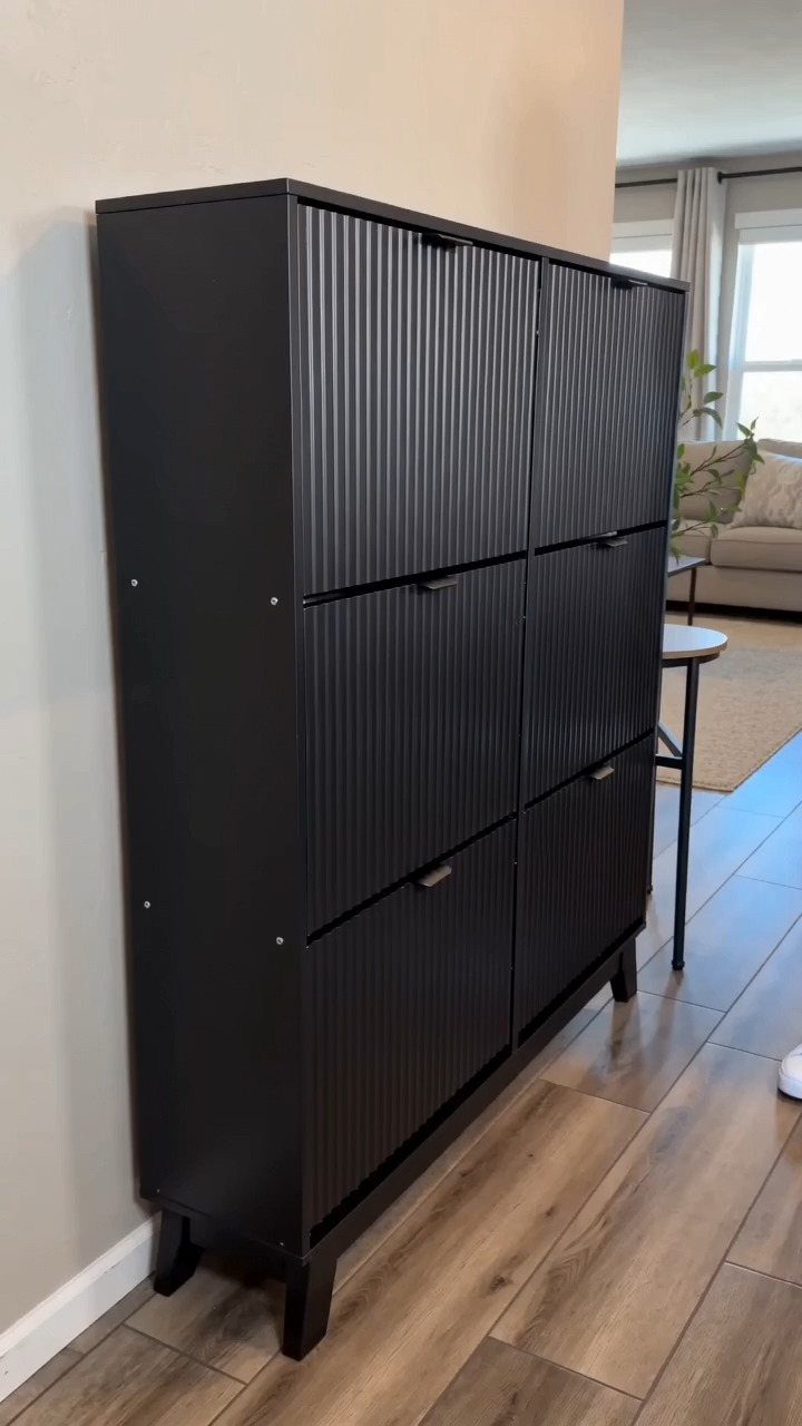 Black fluted shoe storage cabinet with six doors standing on a wooden floor.