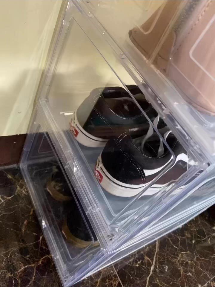 Clear plastic shoe boxes stacked on a marbled floor containing black sneakers.