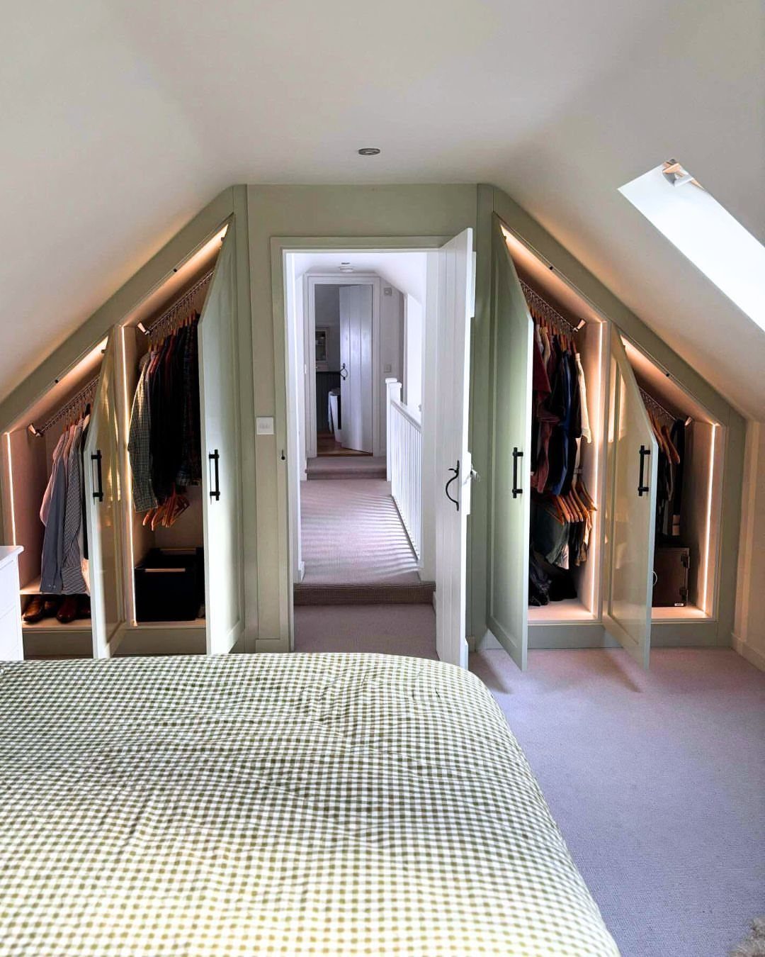 Sage green built-in closets under a slanted ceiling with integrated lighting and hanging clothes.