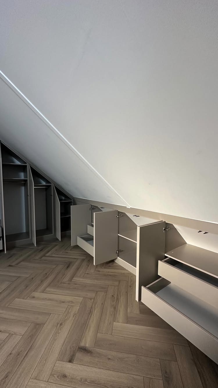 White built-in drawers and cabinets installed under a slanted ceiling with herringbone floors.