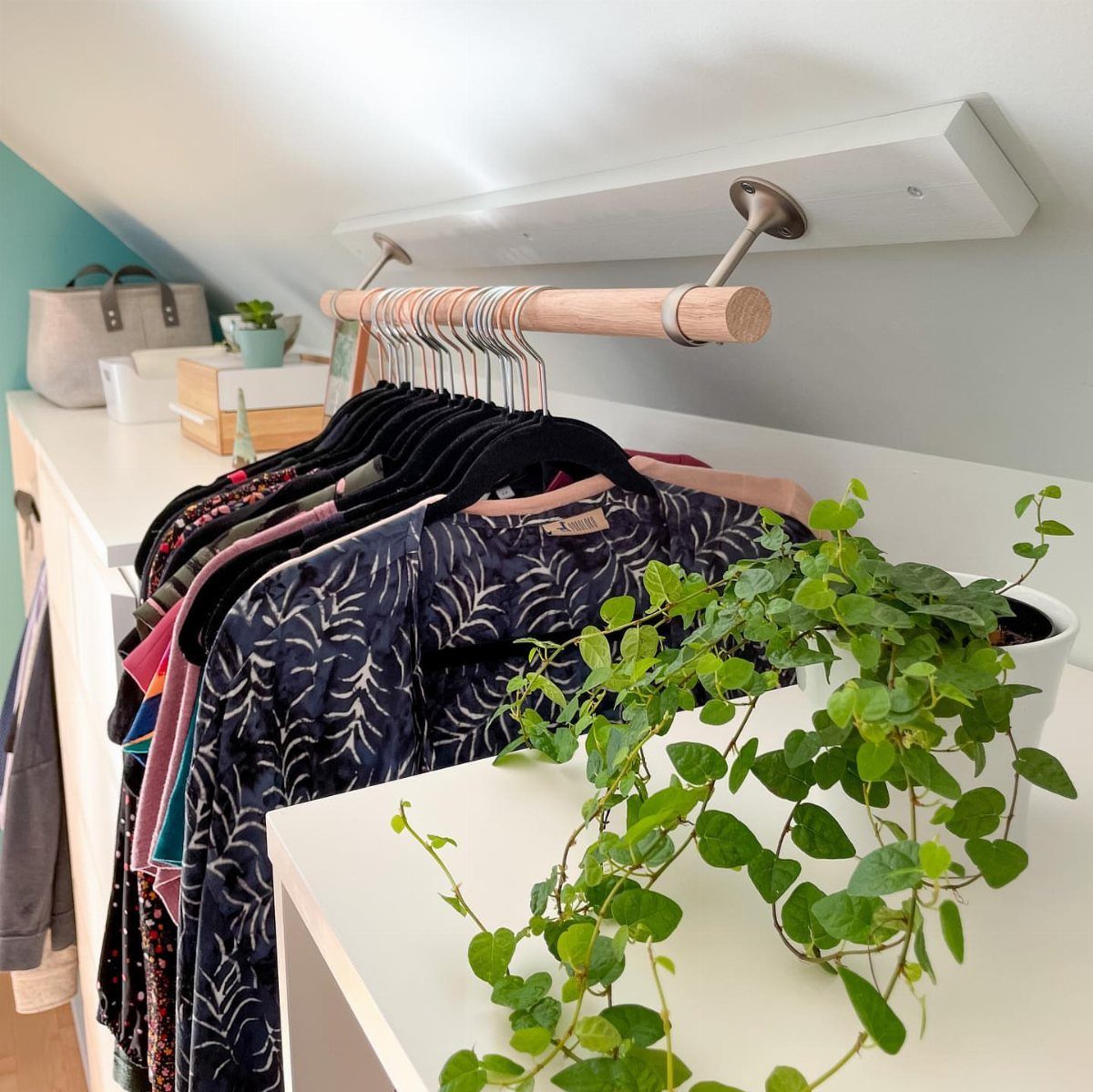 Wooden hanging rod with clothes under a slanted white ceiling with shelving.
