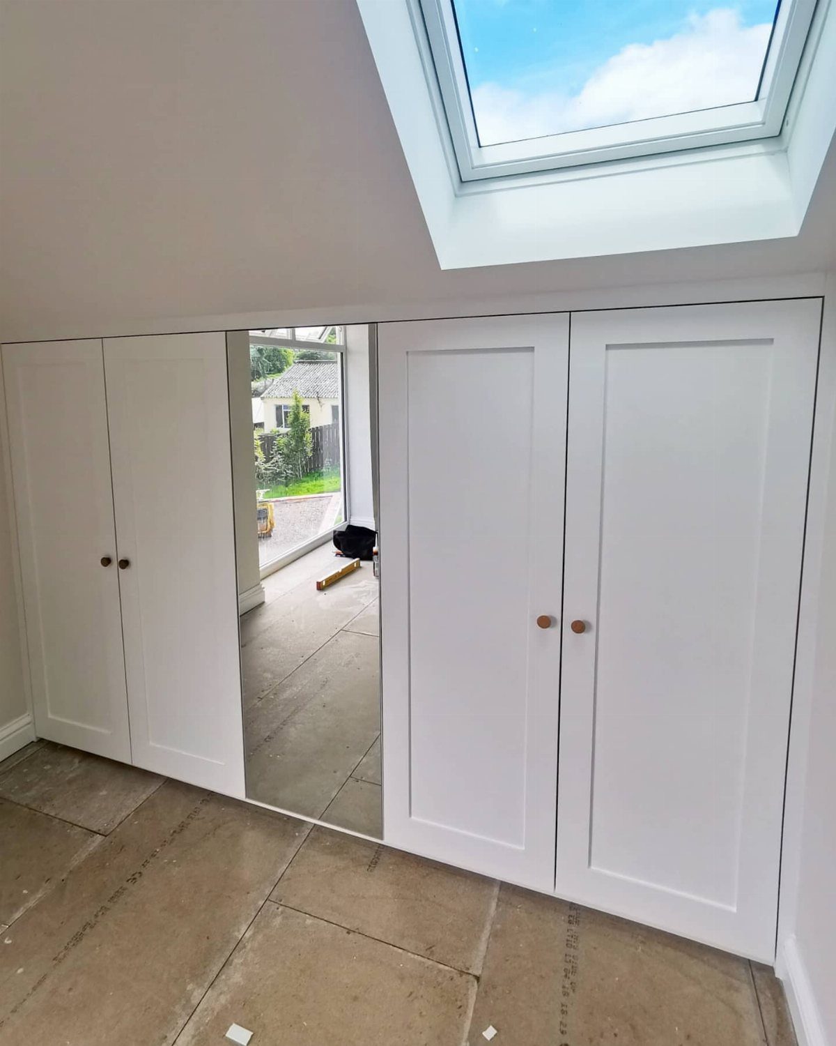 White built-in wardrobes with a mirrored door under a slanted ceiling with skylight.