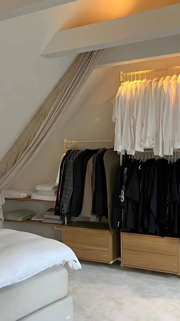 Open closet with hanging clothes and wooden drawers under a slanted bedroom ceiling