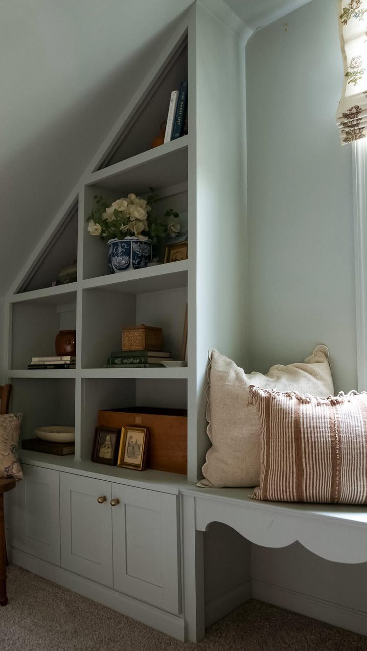 Built-in light grey shelving and cabinets fitted under a slanted ceiling with window seat.