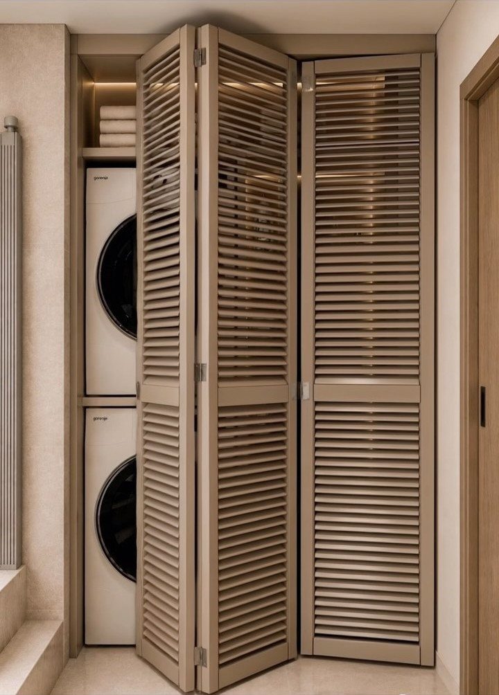 Stacked white washer and dryer behind beige slatted folding doors in a small nook.