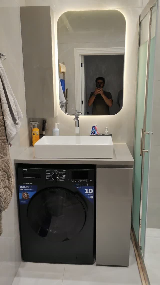 Black washing machine installed under a white bathroom sink and countertop with mirror.