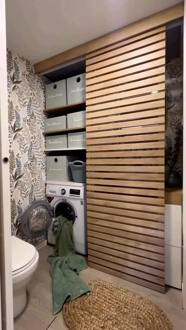 Wooden slatted sliding door partially concealing a washing machine and organized storage bins.