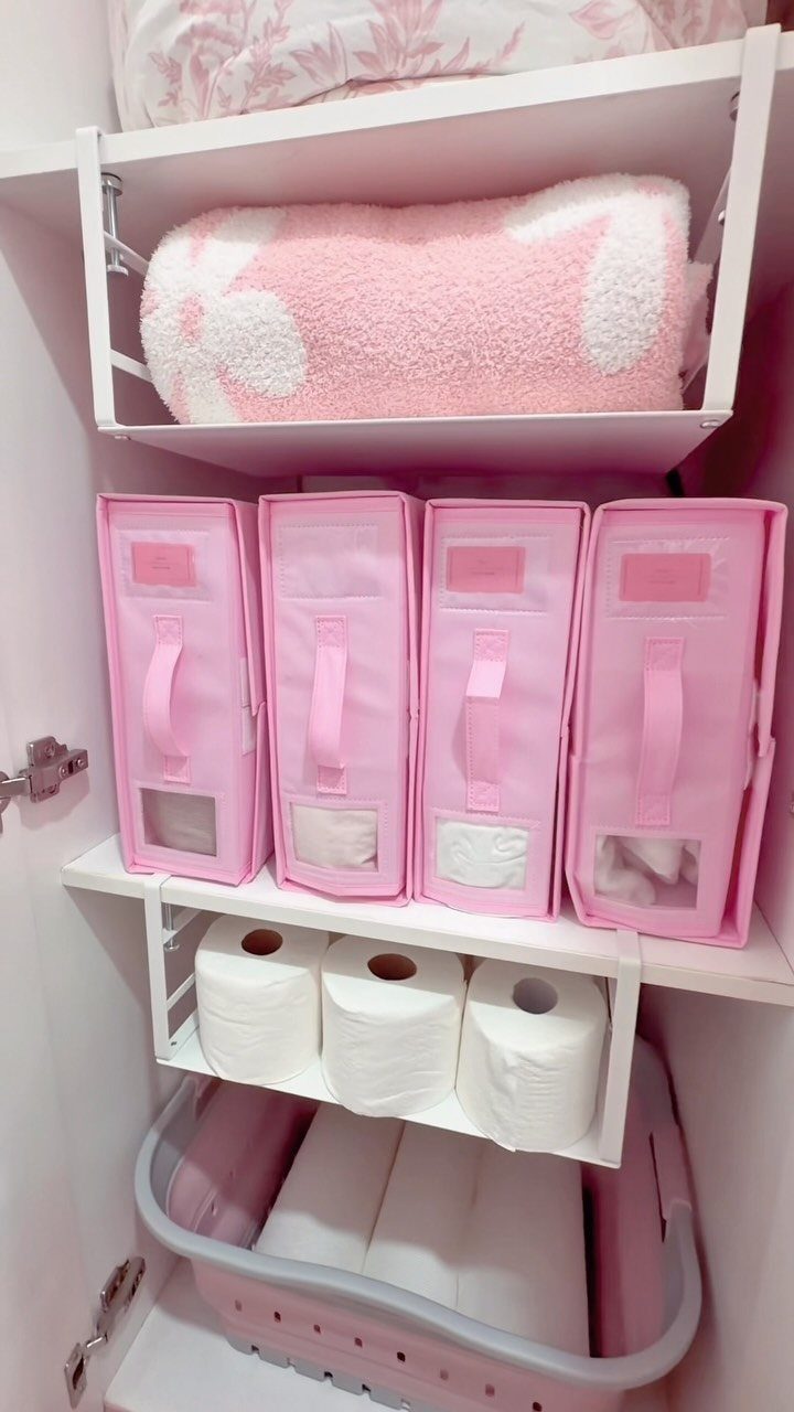 Organized white storage closet with pink bins, towels, and toilet paper rolls.