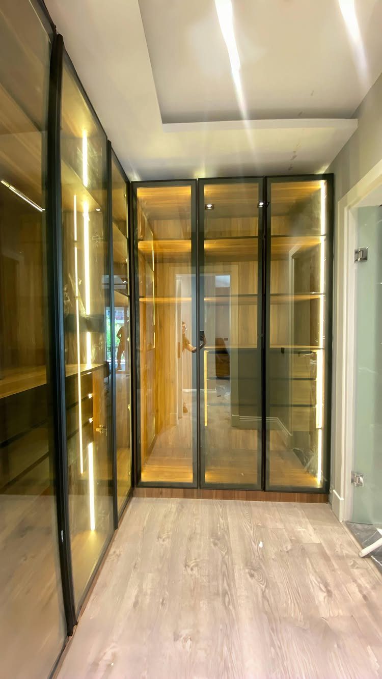 Narrow walk-in closet with glass doors, light wood shelving, and integrated LED lighting.