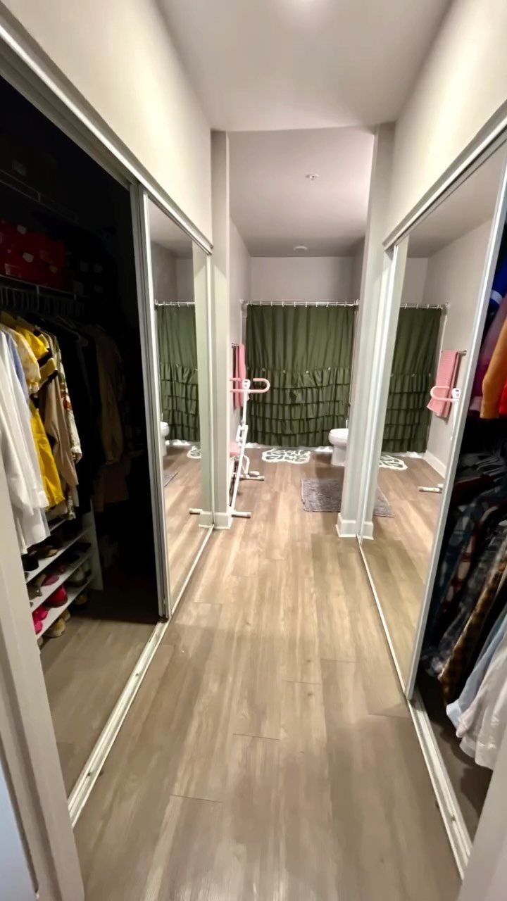 Narrow walk-in closet with mirrored doors, hanging clothes, and light wood flooring.