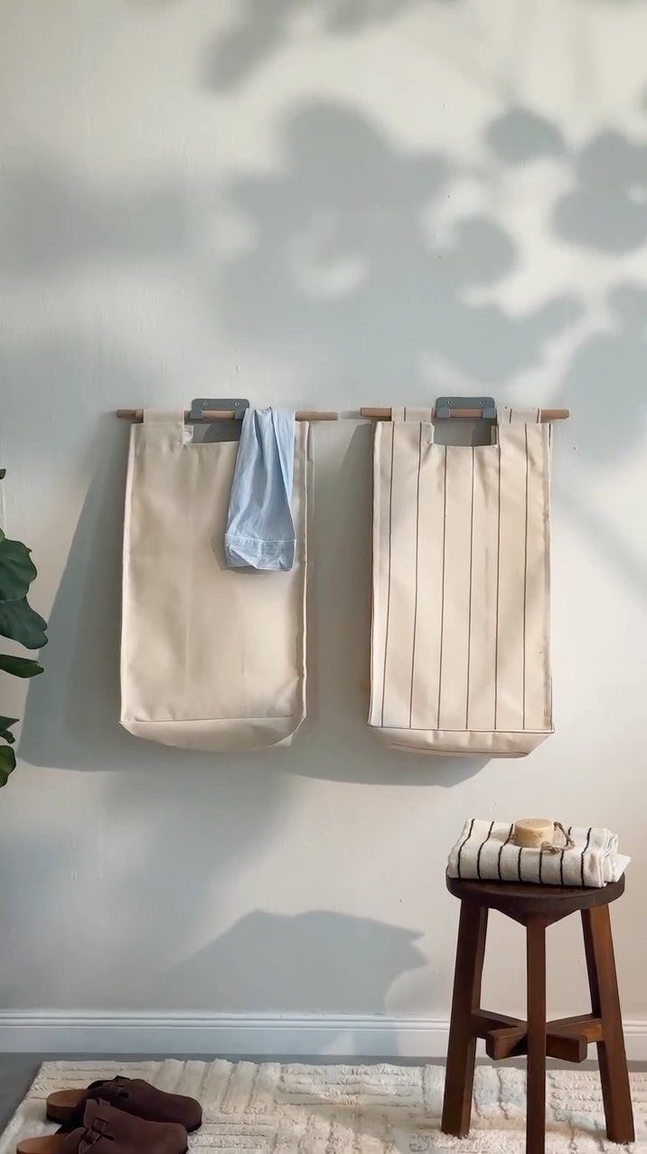 Two beige fabric laundry hampers hanging from wooden rods against a grey wall.
