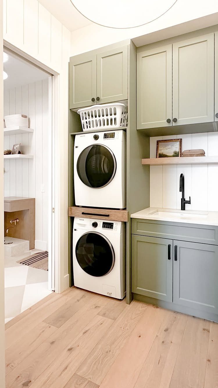 Stacked white washer and dryer surrounded by sage green cabinets and wooden floors.