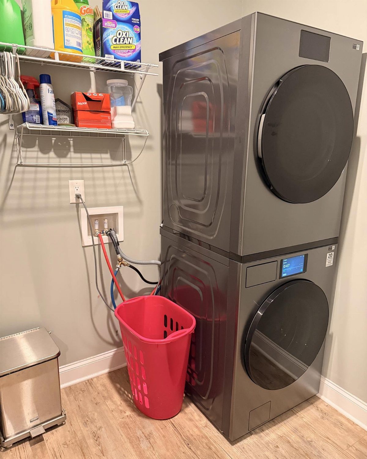 Stacked grey washer and dryer with white wire shelving and red laundry basket.