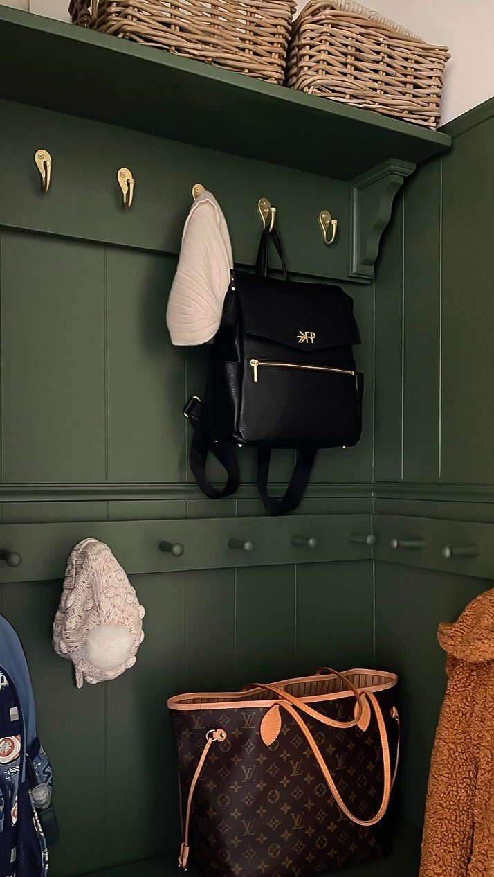 Dark green painted mudroom with gold hooks, wicker baskets, and hanging bags.