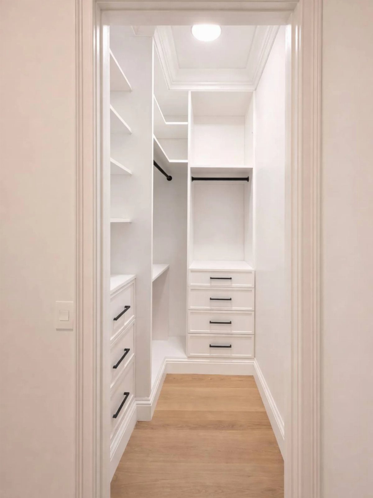 Small white walk-in closet with built-in shelving, drawers, and light wood flooring.