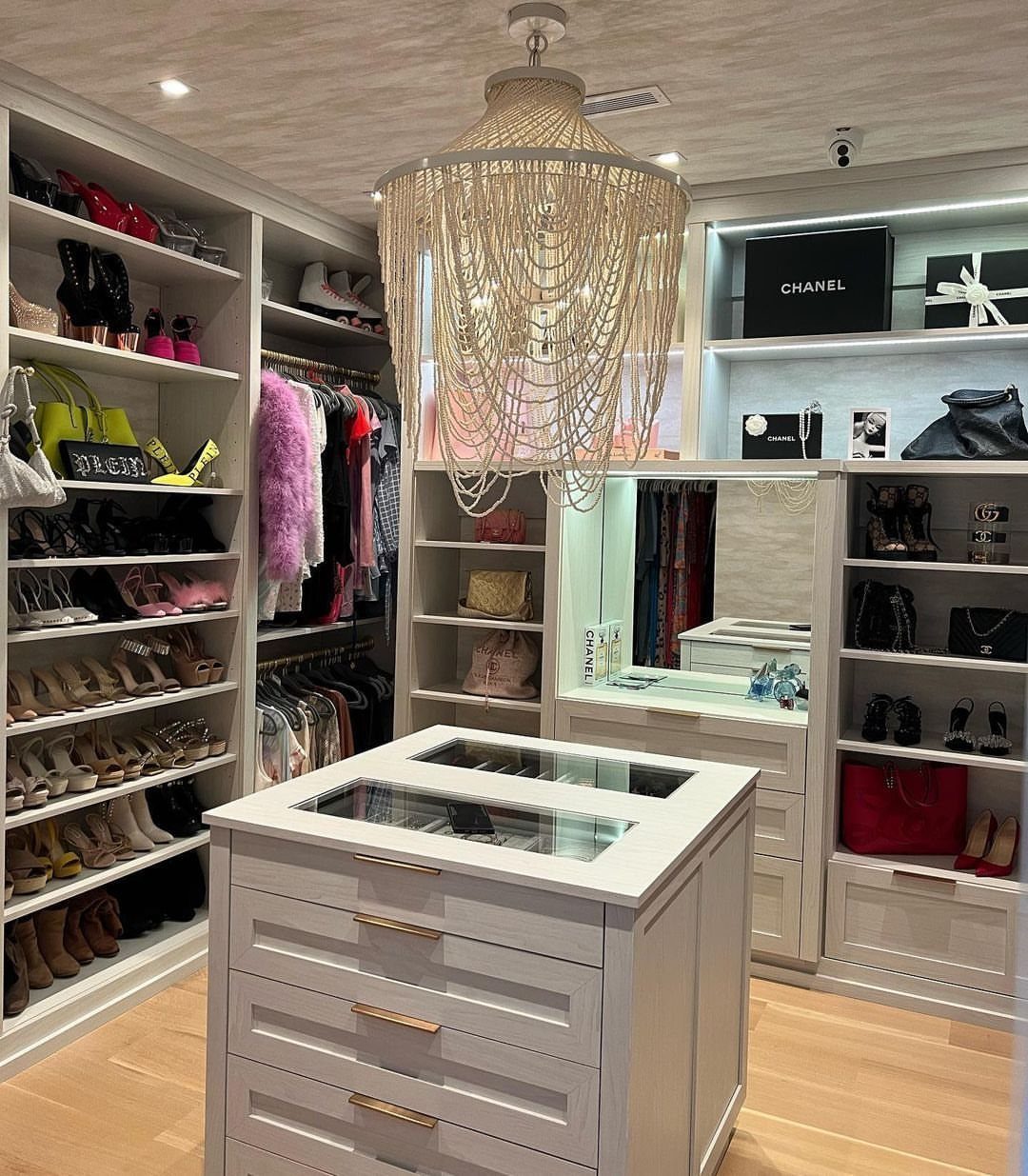 White walk-in closet island with glass top and drawers under a beaded chandelier