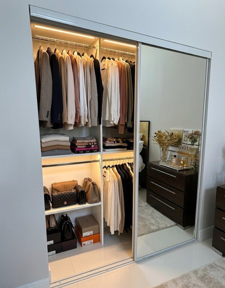 Modern walk-in closet with white shelving and a sliding mirrored door reflecting a dresser.