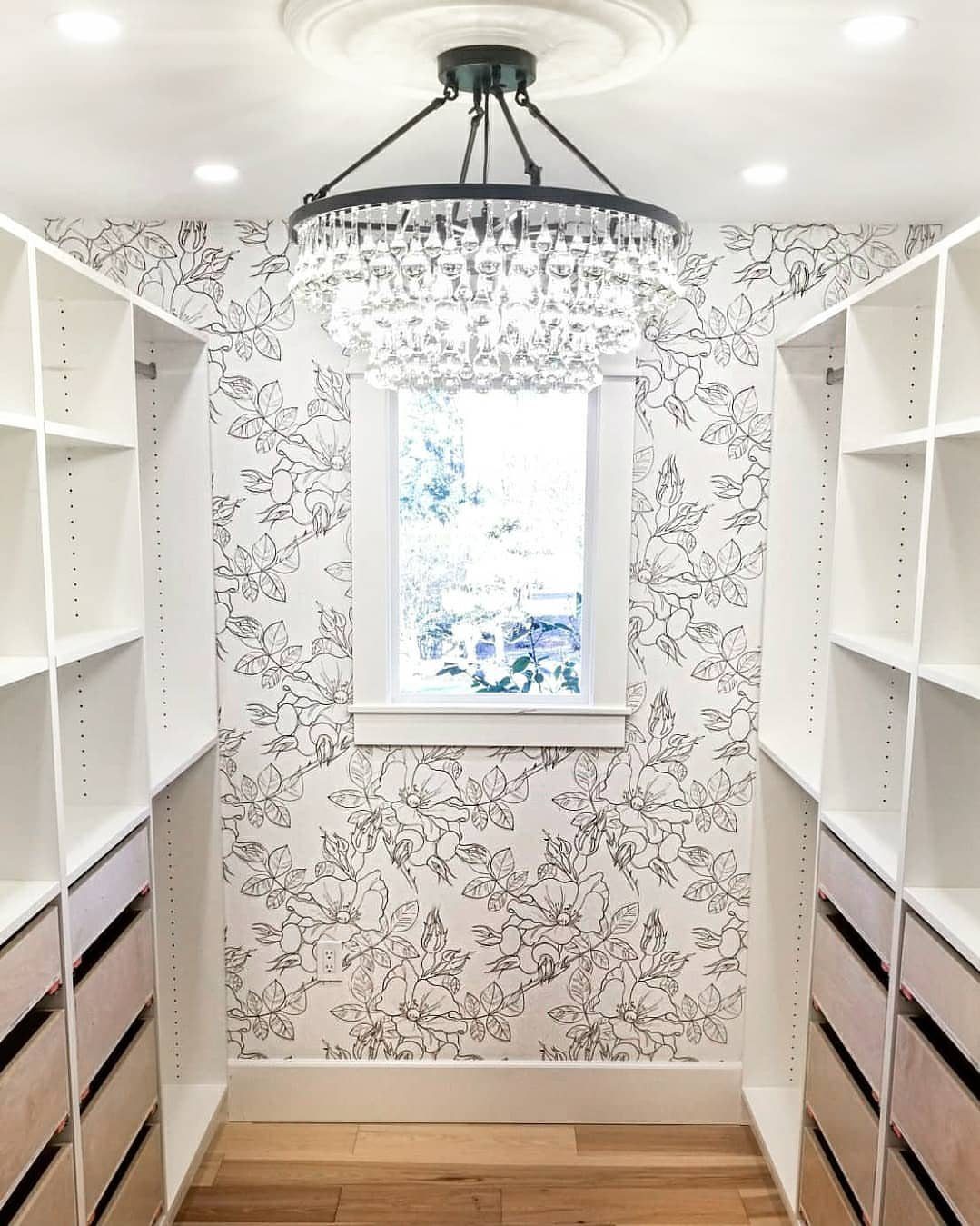 Walk-in closet with black and white floral wallpaper, white shelving, and crystal chandelier.