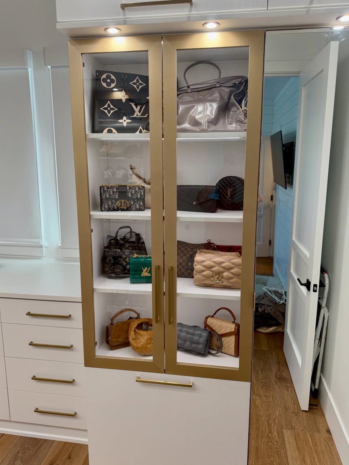 Luxury handbags in a gold-framed cabinet illuminated by overhead puck lights.