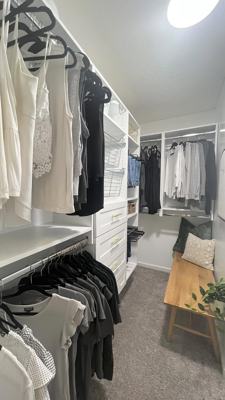 White modular closet system with hanging clothes and a wooden bench on grey carpet.