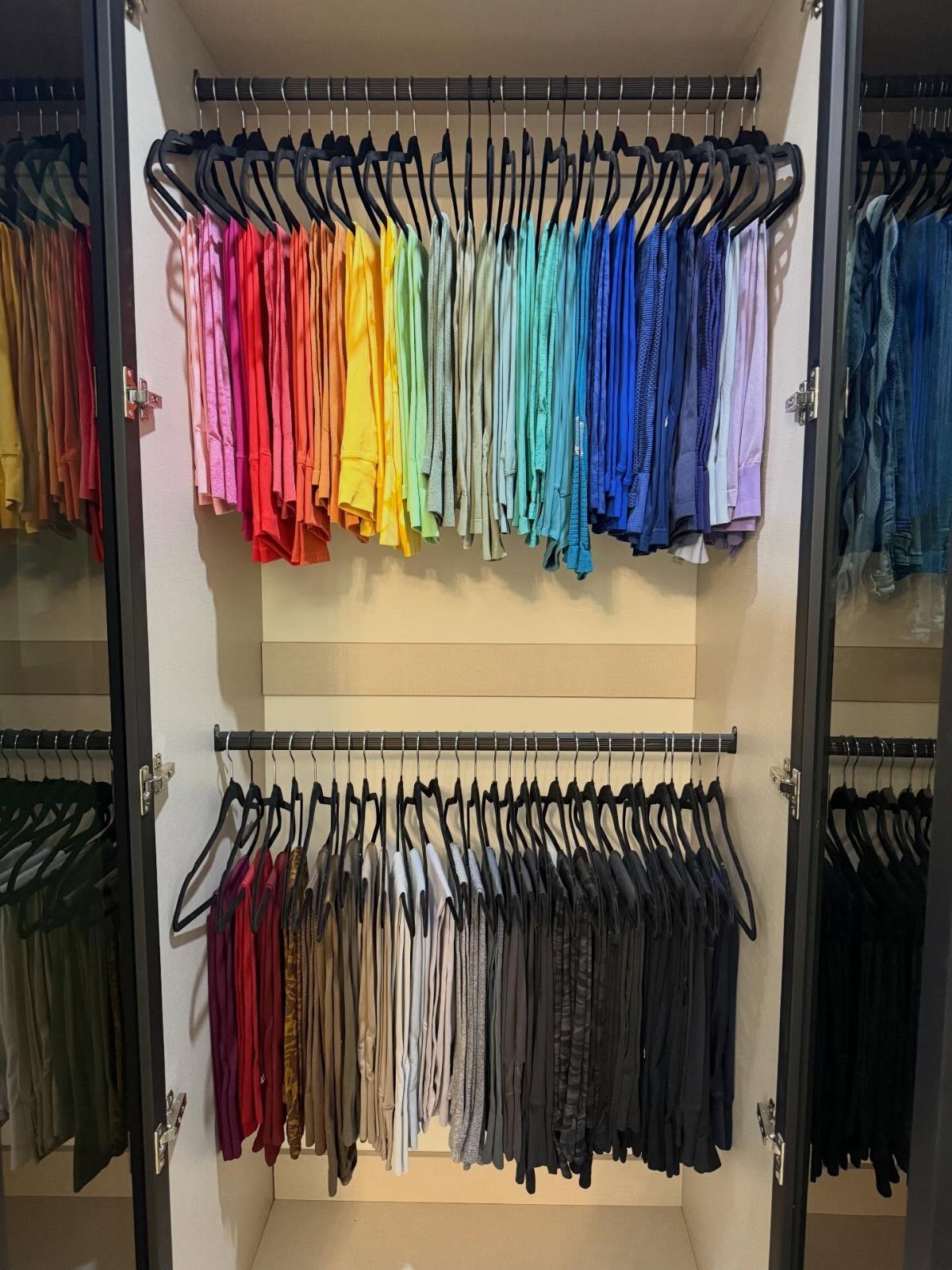 Shirts arranged in rainbow and neutral color gradients on black hangers in a closet.