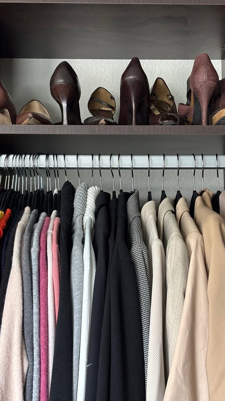 Slim white hangers holding a variety of neutral colored clothing under a shoe shelf