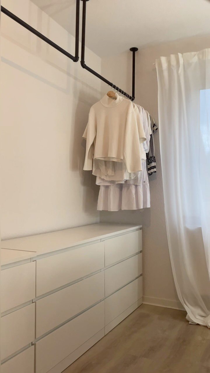 White clothing hanging from a black industrial pipe rod above white drawers.