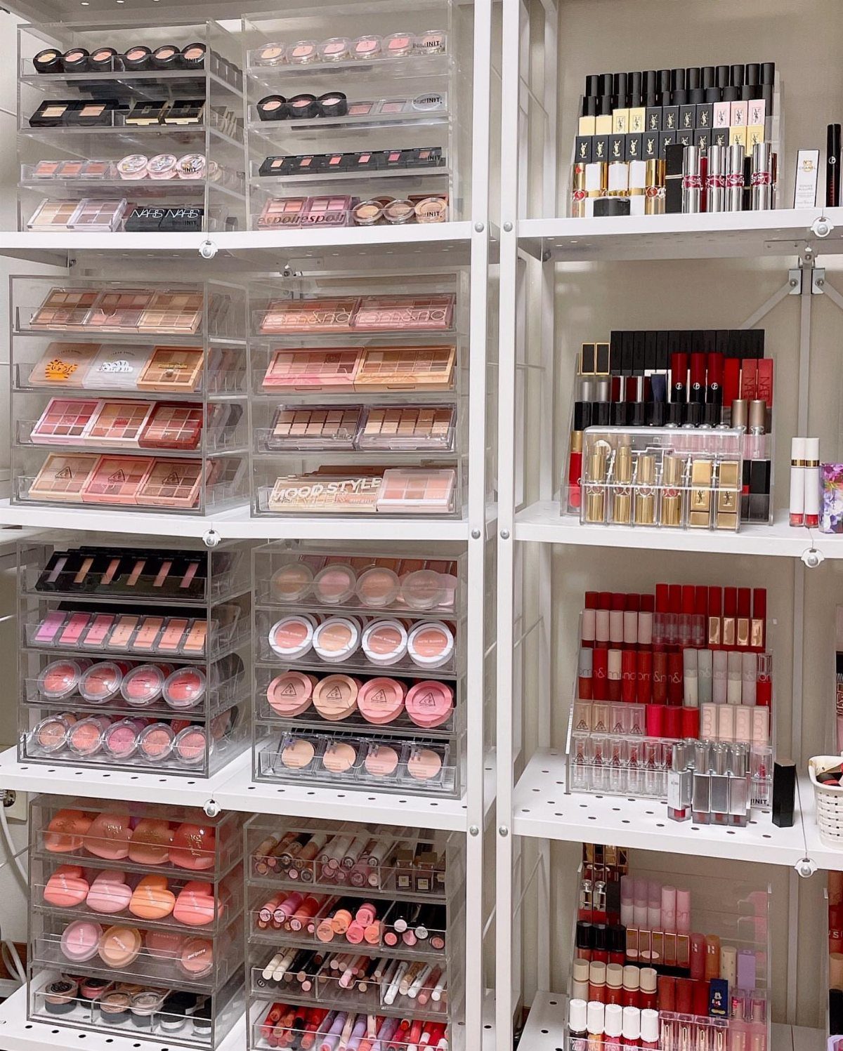 Acrylic makeup organizers filled with cosmetics on white metal shelving units.