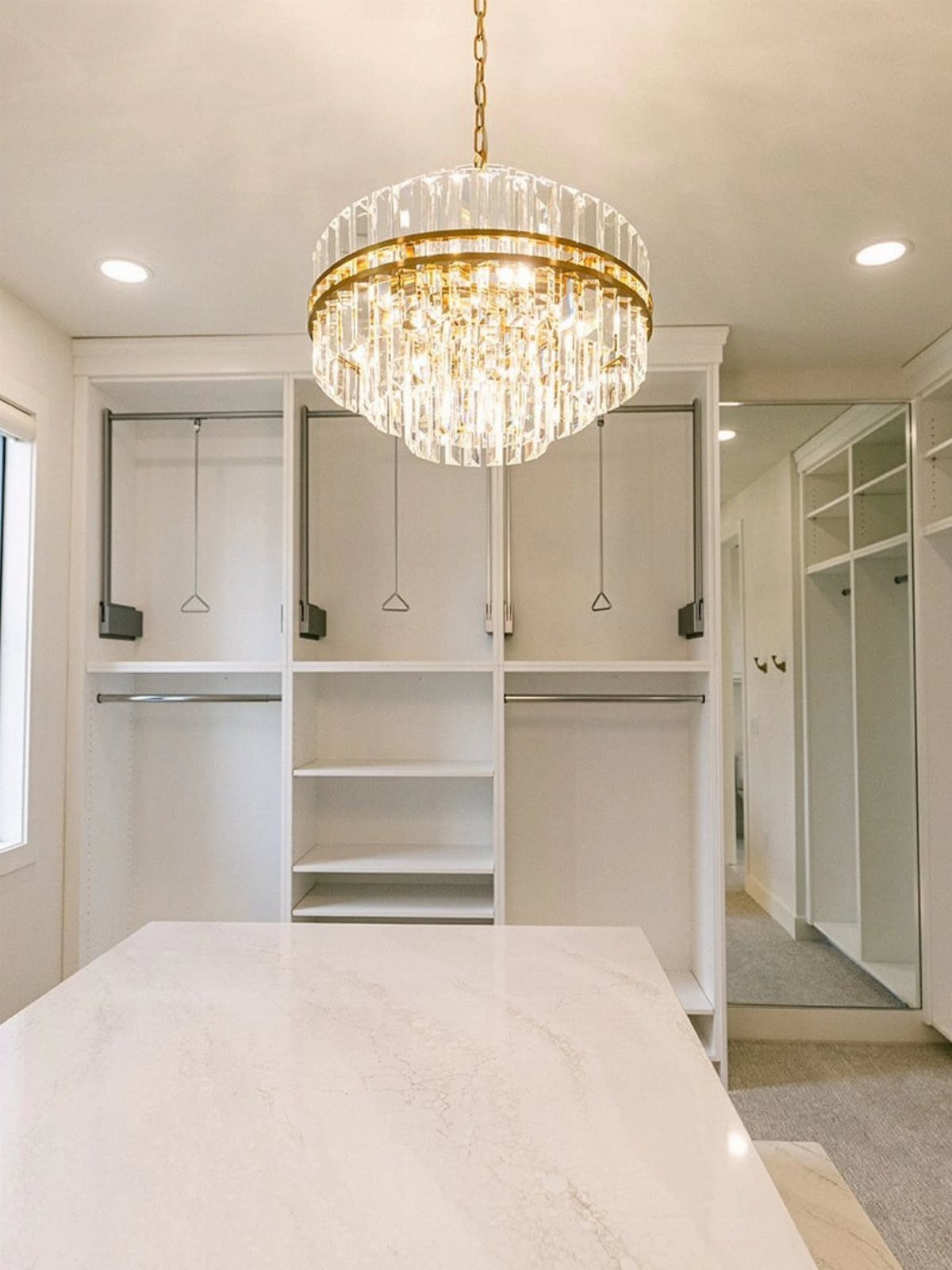 Crystal chandelier hanging in a white walk-in closet with marble countertops and shelving.