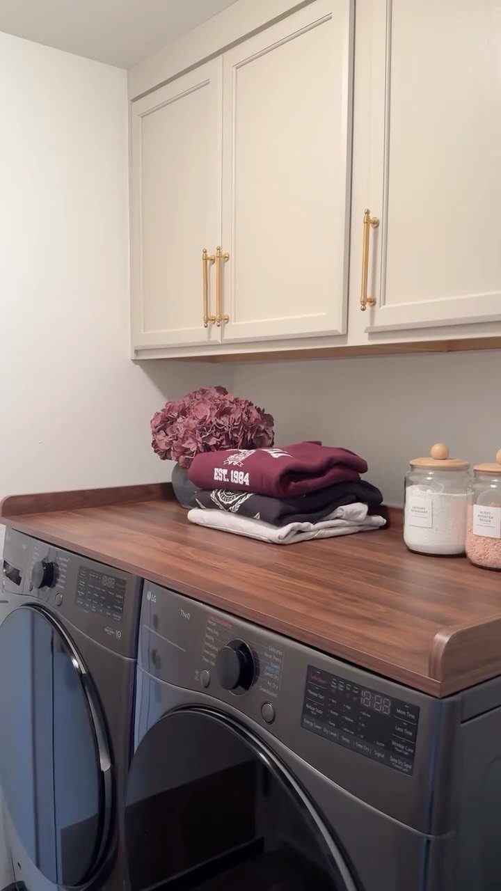 Wooden countertop over a grey washer and dryer with folded laundry and jars.
