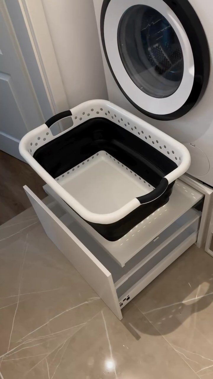 White washing machine on a white storage stand with a pull-out laundry basket drawer